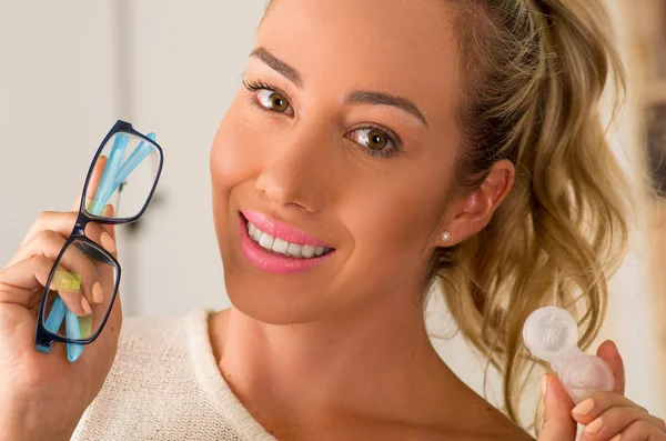 Mujer rubia joven sonriente sosteniendo la caja de lentes de contacto en la mano y sosteniendo en su otra mano unas gafas azules sobre un fondo borroso. — Foto de Stock