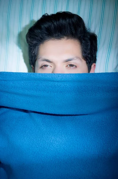 Close up of a handsome young man in bed with eyes opened suffering insomnia and sleep disorder thinking about his problem, covered half of his face with a blue blanket, above view