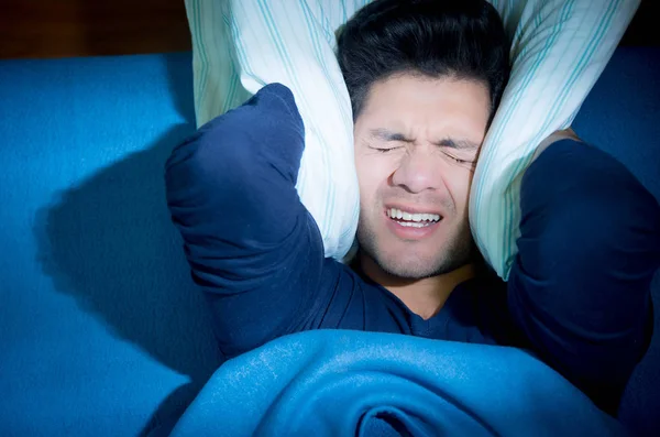 Красивий молодий чоловік у ліжку страждає безсонням і розладом сну, прикриваючи вуха подушкою, зверху — стокове фото