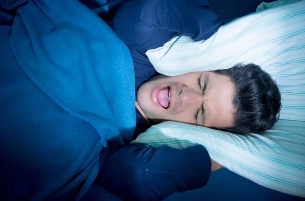 Joven guapo en la cama sufriendo insomnio y trastorno del sueño, cubriéndose los oídos con una almohada —  Fotos de Stock
