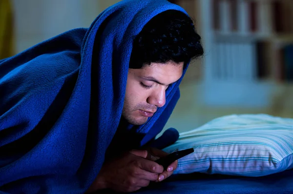 Media społecznościowe-addict mężczyzn na łóżku nie spać, bo grać inteligentny telefon, obejmujące głowę z niebieski koc, tło pokoju — Zdjęcie stockowe