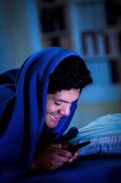 男人在床上不睡觉，因为社交媒体瘾君子玩智能手机，涵盖抬起头来，一条蓝色毯子房间背景 — 图库照片