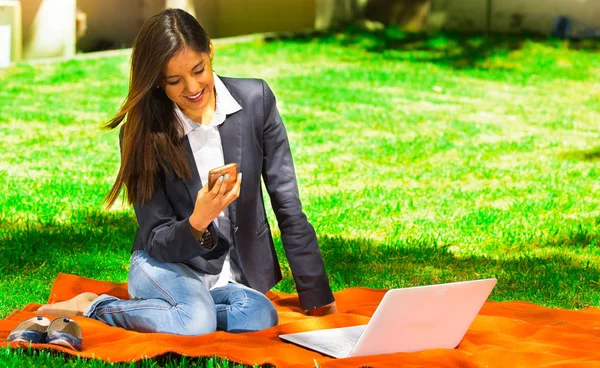 Chica joven y feliz con portátil en el parque usando su teléfono celular — Foto de Stock