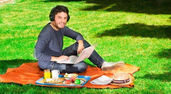 Gelukkig jongeman picknick in het park genieten terwijl hij gebruikt zijn computer een koptelefoon dragen — Stockfoto