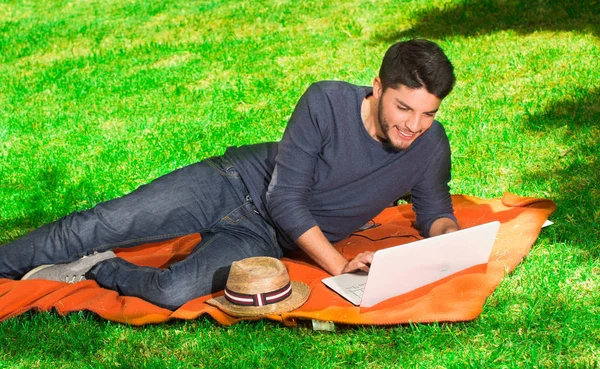 Schöner junger Mann im Park, der mit einem Laptop auf dem Rasen sitzt — Stockfoto