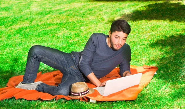 Guapo joven se ve sorprendido en el parque sentado en la hierba con un ordenador portátil —  Fotos de Stock