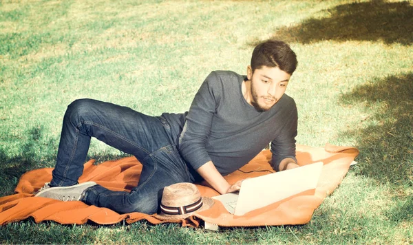 Красивый молодой человек выглядит удивленным в парке сидя на траве с ноутбуком, винтажный эффект — стоковое фото