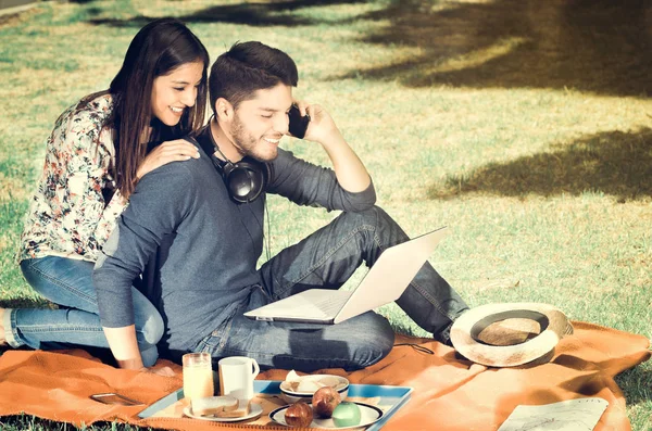 Jovem casal feliz curtindo piquenique no parque enquanto o cara usa seu celular, efeito vintage — Fotografia de Stock