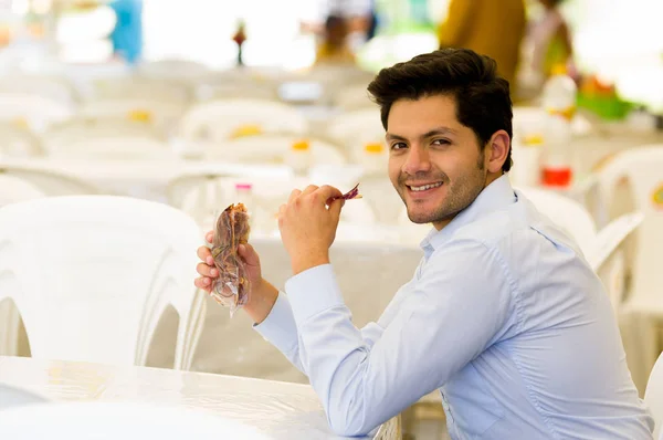 Bonito e sorridente jovem empresário comendo uma deliciosa fatia de batata doce frita dentro de um saco de plástico em um mercado público — Fotografia de Stock