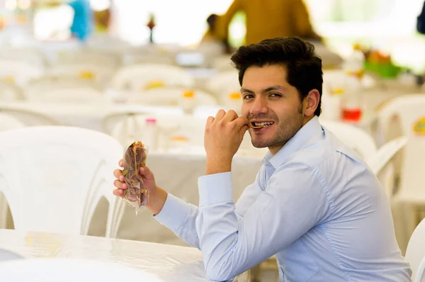 Bonito e sorridente jovem empresário comendo uma deliciosa fatia de batata doce frita dentro de um saco de plástico em um mercado público — Fotografia de Stock