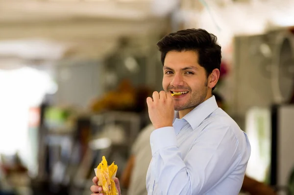 Bonito jovem empresário comendo uma deliciosa fatia de banana frita dentro de um saco de plástico em um mercado público — Fotografia de Stock