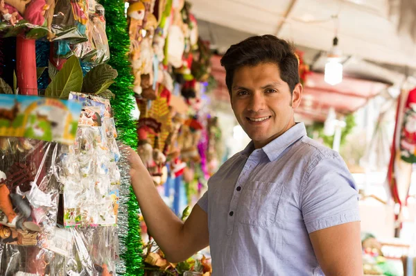 Красивый молодой человек смотрит красивое и красочное украшение рождественской елки на рынке — стоковое фото