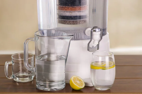 Filter system of water purifier with two glasses of water one filled until middle with a lemon inside and an empty pitcher on wooden table — Stock Photo, Image