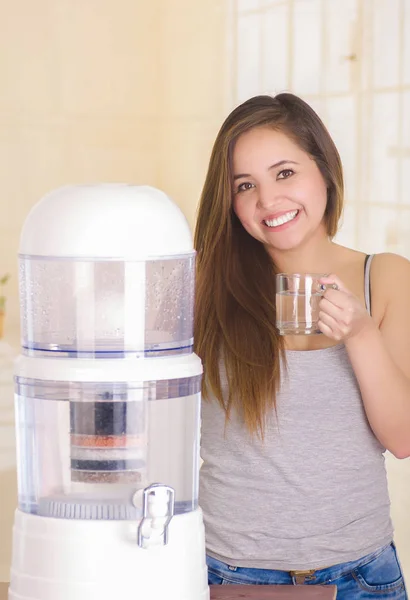 Bella donna sorridente che tiene un bicchiere d'acqua dietro di un sistema di filtro di depuratore d'acqua su uno sfondo di cucina — Foto Stock