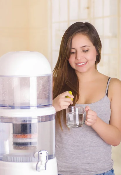Bella donna sorridente aggiungendo un po 'di succo di limone nel bicchiere d'acqua, sistema di filtraggio del depuratore d'acqua su uno sfondo della cucina — Foto Stock