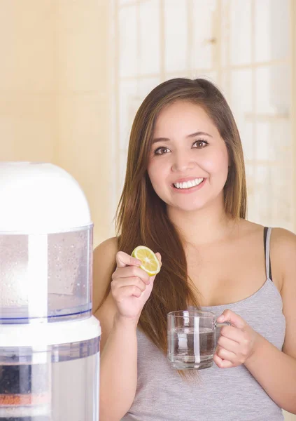 Bella donna sorridente che tiene un bicchiere d'acqua dietro di un sistema di filtro di depuratore d'acqua su uno sfondo di cucina — Foto Stock