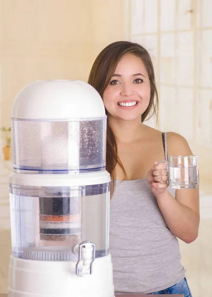 Красивая улыбающаяся женщина, держащая стакан воды за фильтрующей системой очистителя воды на кухонном фоне — стоковое фото