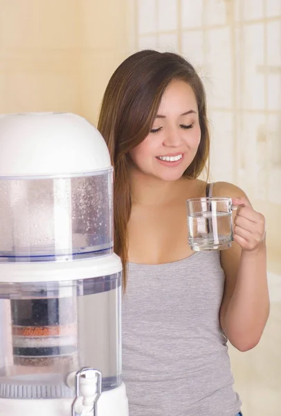キッチンの背景に浄水器のフィルター システムの背後にある水のガラスを保持して笑顔美人 — ストック写真