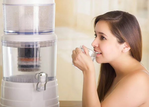 Zbliżenie piękna kobieta wypija szklankę wody z filtra system oczyszczania wody na tle kuchnia — Zdjęcie stockowe