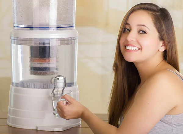 Close up van een mooie lachende vrouw vullen van een glas water, met een systeem van de filter van water purifier op de achtergrond van een keuken — Stockfoto