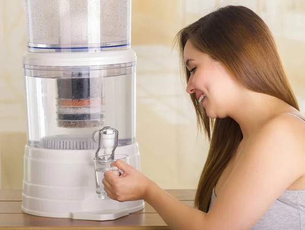 Закрыть глаза на красивую улыбчивую женщину, наполняющую стакан водой, с системой фильтрации очистителя воды на кухонном фоне — стоковое фото