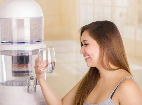 Красивая улыбающаяся женщина, держащая стакан воды, с системой фильтрации очистителя воды на кухонном фоне — стоковое фото