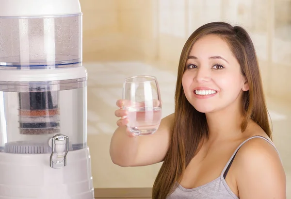 Mooie lachende vrouw met een glas water, met een systeem van de filter van water purifier op de achtergrond van een keuken — Stockfoto