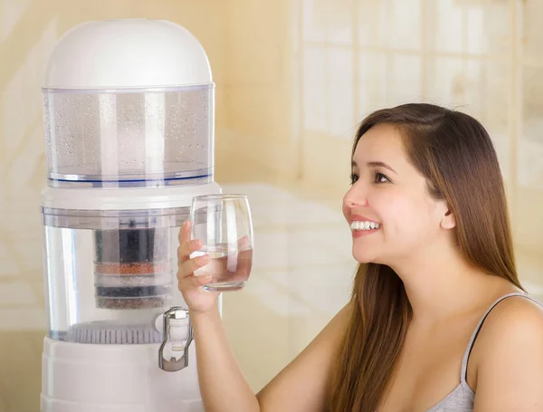 Piękna kobieta uśmiechający się trzymając szklankę wody z filtra system oczyszczania wody na tle kuchnia — Zdjęcie stockowe