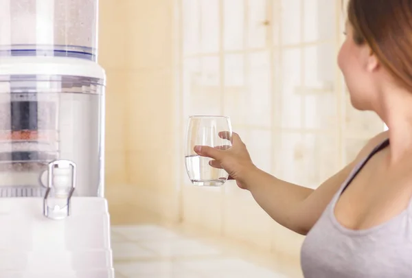 Beautiful smiling woman holding a glass of water, with a filter system of water purifier on a kitchen background — Stock Photo, Image