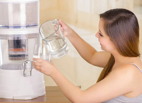 Beautiful woman holding a glass of water in one hand and a pitcher of water in her other hand, with a filter system of water purifier on a kitchen background — Stock Photo, Image