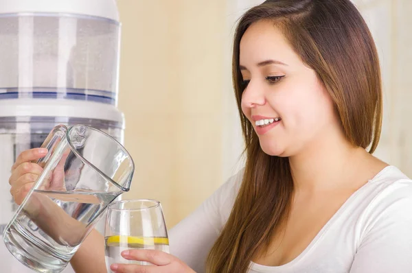 Beautiful woman holding a glass of water in one hand and a pitcher of water in her other hand, with a filter system of water purifier on a kitchen background — Stock Photo, Image