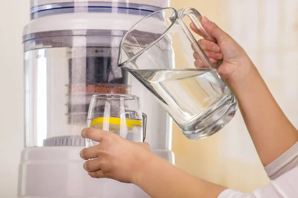 Close up of a woman holding a glass of water in one hand and a pitcher of water in her other hand, with a filter system of water purifier on a kitchen background — Stock Photo, Image