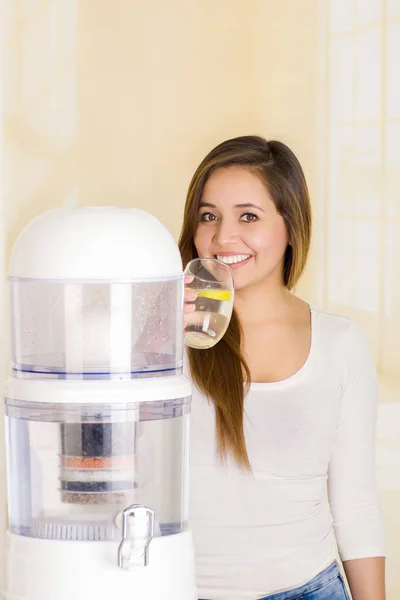 Bella donna che beve un bicchiere d'acqua con un sistema di filtraggio del depuratore d'acqua su uno sfondo della cucina — Foto Stock
