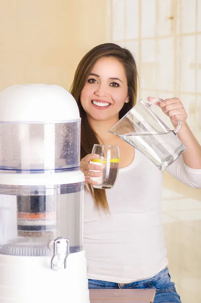 Mooie vrouw met een glas water in één hand en een kruik water in haar andere hand, met een systeem van de filter van water purifier op de achtergrond van een keuken — Stockfoto