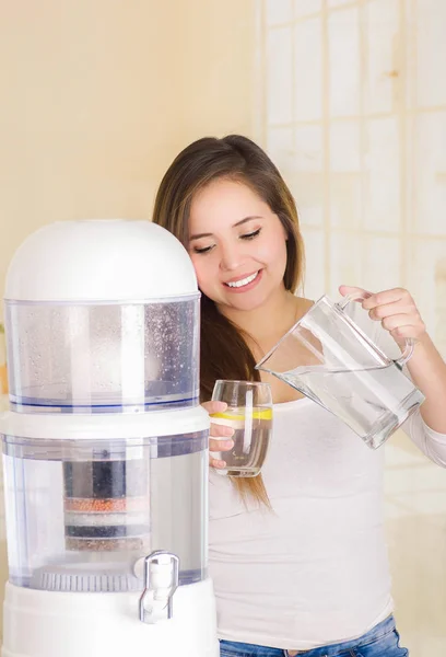 Mooie vrouw met een glas water in één hand en een kruik water in haar andere hand, met een systeem van de filter van water purifier op de achtergrond van een keuken — Stockfoto