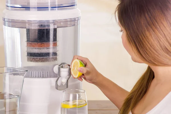 Mooie vrouw met een gesneden citroen in haar hand met een systeem van de filter van water purifier op de achtergrond van een keuken — Stockfoto