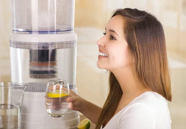 Hermosa mujer sonriente sosteniendo un limón cortado en su mano con un sistema de filtro de purificador de agua sobre un fondo de cocina —  Fotos de Stock