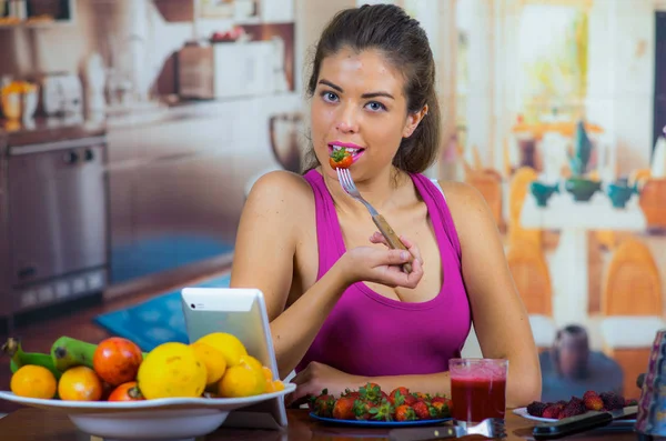 Giovane donna che indossa top rosa godendo di colazione sana, mangiare fragole e sorridente, sfondo cucina di casa — Foto Stock