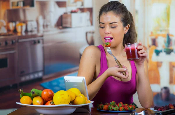 Jeune femme vêtue d'un haut rose savourant un petit déjeuner sain, mangeant des fraises et souriant, fond de cuisine maison — Photo