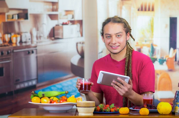 Giovane uomo con i timori di godere di una colazione sana, mangiare frutta, bere frullato e sorridere con tablet, sfondo cucina di casa — Foto Stock