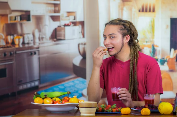 Giovane uomo con i dreads godendo la prima colazione sana, mangiare frutta, bere frullato e sorridente, sfondo cucina di casa — Foto Stock