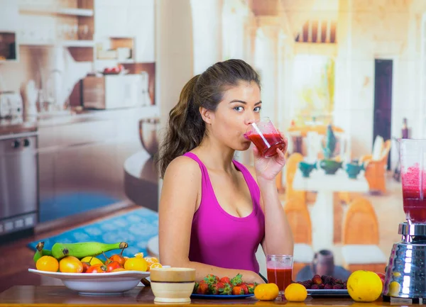 Giovane donna che indossa top rosa godendo di colazione sana, mangiare frutta, bere frullato e sorridente, sfondo cucina di casa — Foto Stock