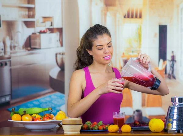 Giovane donna che indossa top rosa godendo di colazione sana, mangiare frutta, utilizzando frullatore preparazione frullato e sorridente, sfondo cucina di casa — Foto Stock