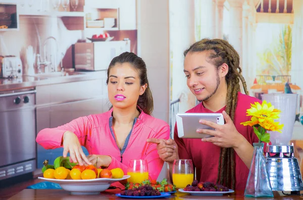 Pareja hispana joven y saludable disfrutando del desayuno juntos, compartiendo frutas y sonriendo mientras miran la pantalla de la tableta, fondo de cocina casera — Foto de Stock