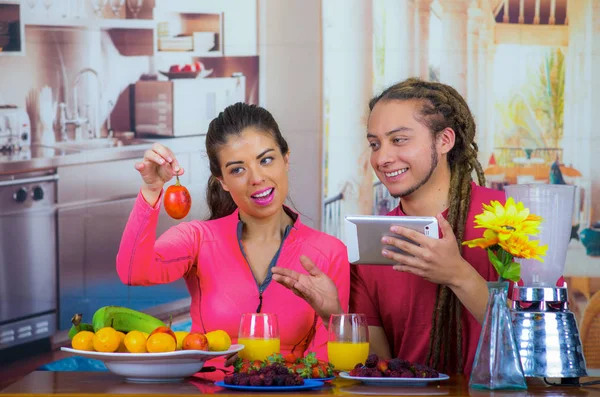 Pareja hispana joven y saludable disfrutando del desayuno juntos, compartiendo frutas y sonriendo mientras miran la pantalla de la tableta, fondo de cocina casera — Foto de Stock