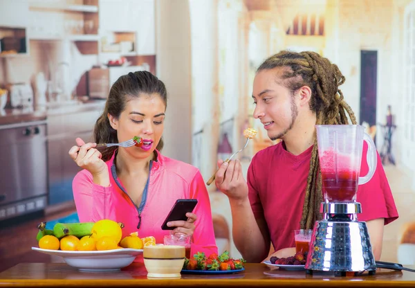 Ispanico giovane coppia sana godendo la colazione insieme, la condivisione di frutta, fissando lo schermo mobile e sorridente, sfondo cucina di casa — Foto Stock