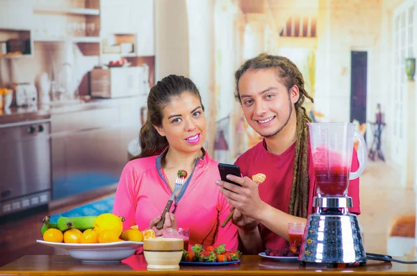 Hispanic jonge gezonde paar genieten van ontbijt samen, delen van vruchten, drinken smoothie en lachende, huis keuken achtergrond — Stockfoto