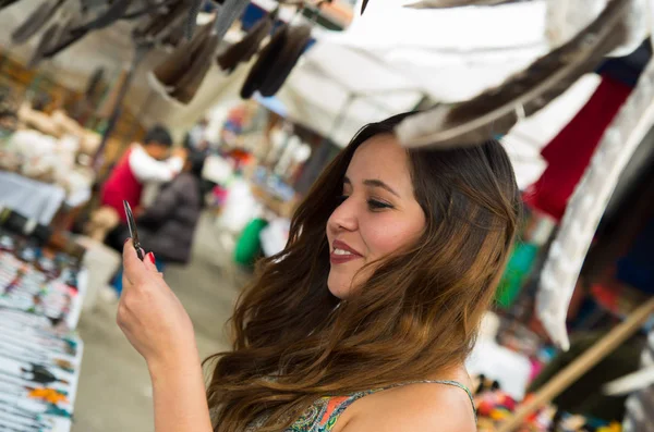 Hermosa mujer mirando algo en la ropa tradicional andina y artesanías con una pluma borrosa en el frente, fondo del mercado — Foto de Stock