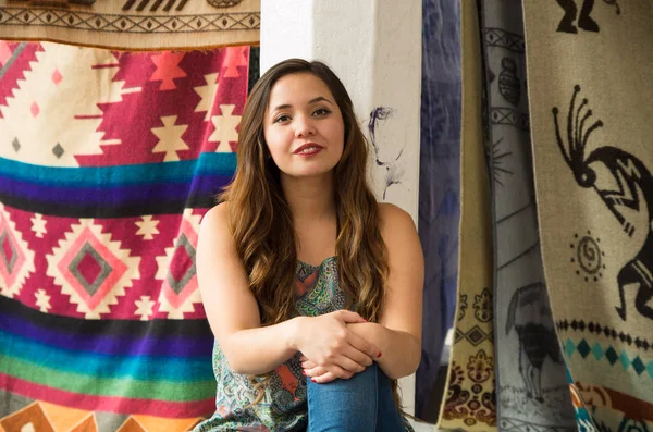 Mooie jonge vrouw met beide handen over haar knie, Andes traditionele kleding textielgarens en geweven door hand in wol, kleurrijke stoffen achtergrond — Stockfoto