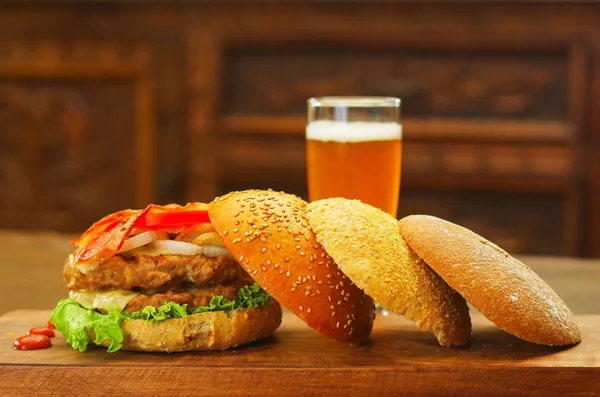 Deliciosas hamburguesas con carne de res, tocino, cebolla, tomate, lechuga y queso con un vaso de cerveza detrás con tres piezas de pan sobre tabla de madera —  Fotos de Stock